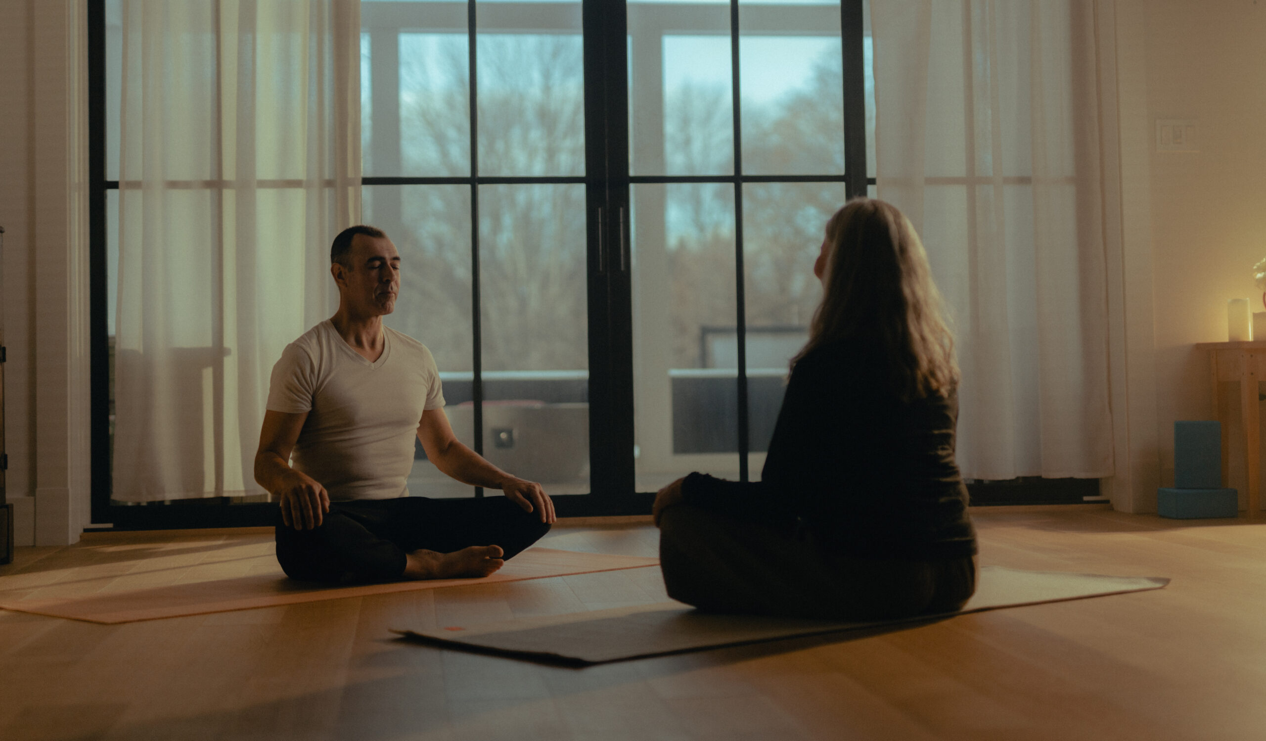 A man and a woman sit across from one another on yoga mats in lotus position.