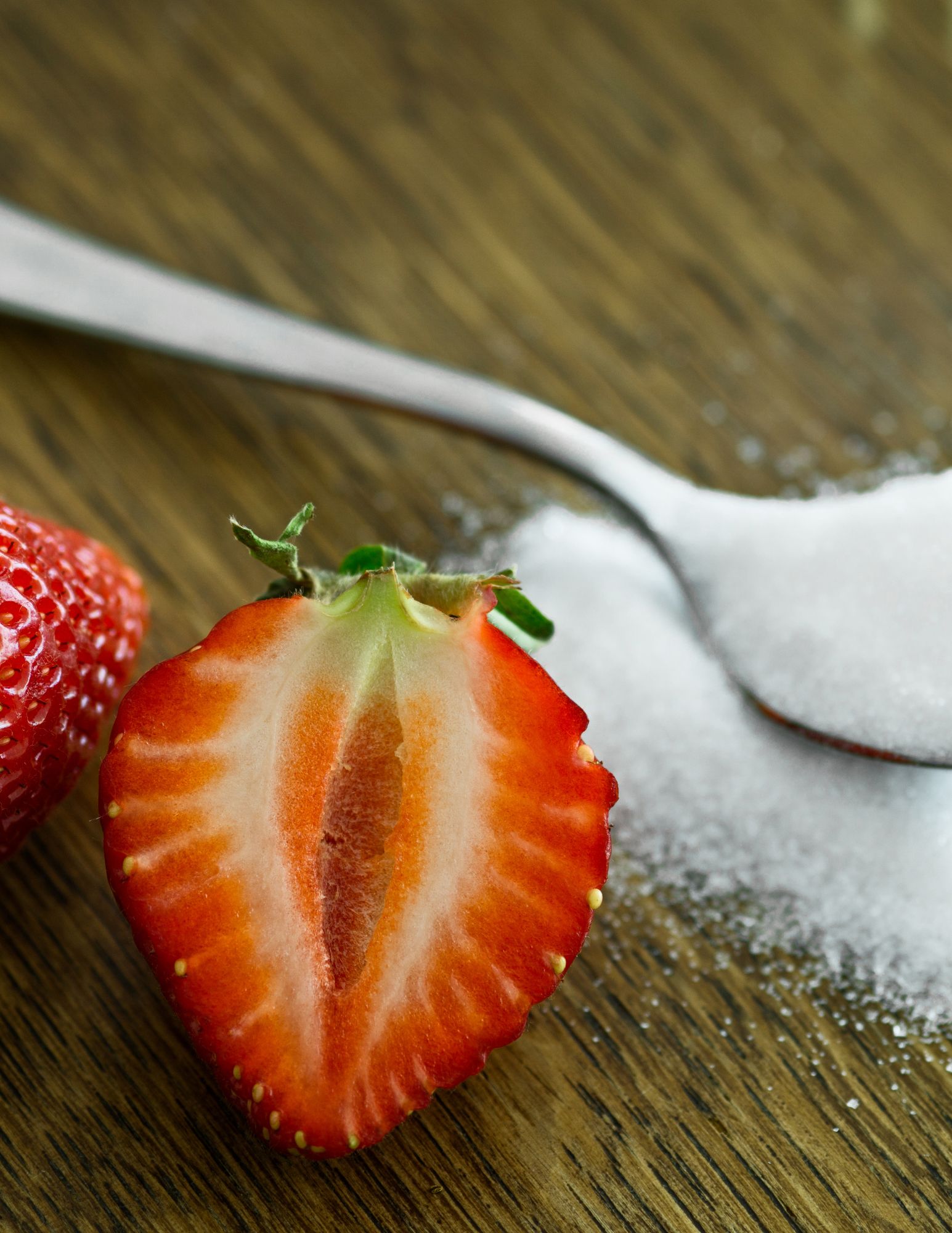 a strawberry halved in the foreground and a teaspoon of sugar in background