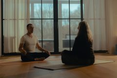 A man and a woman sit across from one another on yoga mats in lotus position.